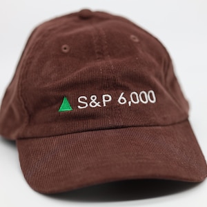 May include: A brown corduroy baseball cap featuring the text "S&P 6,000" embroidered in white, with a green upward-pointing triangle. The cap has a curved peak and a rounded crown.