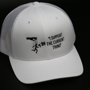 Könnte beinhalten: Weiße Trucker-Kappe mit schwarzem Netzrücken. Die Kappe hat ein schwarzes Motiv einer Marionette mit einem Megaphon und dem Text "I support the current thing".