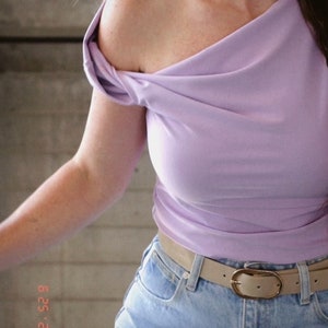 May include: A woman wearing a lavender off-the-shoulder top with a knotted detail on the shoulder. She is wearing a light brown belt with a gold buckle over a pair of light blue jeans.