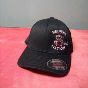 Peut inclure: Casquette de baseball noire avec le texte "REDRUM MC NATION" brodé en blanc et rouge. La casquette a une visière incurvée et une étiquette Flexfit. Taille S/M.
