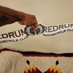 Puede incluir: Dos pegatinas blancas con el texto "REDRUM MOTORCYCLE CLUB" en letras marrón y gris. Las pegatinas presentan un tocado nativo americano con dos calaveras en el centro.
