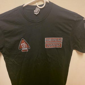 May include: A black short-sleeve t-shirt featuring a red and white triangular patch with the text 13 1/2 on the chest and both sleeves. The chest also displays a rectangular red and white patch with the text REDRUM NATION in bold capital letters.