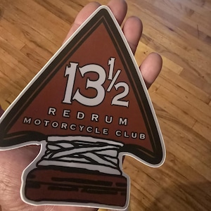 May include: A sticker featuring a brown arrowhead design with the text "13 1/2 REDRUM MOTORCYCLE CLUB" in white. The sticker has a white border and a stylized base with white wrapping.