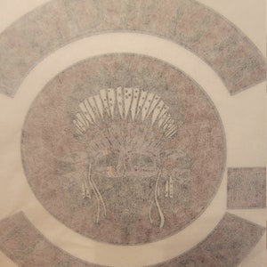 Peut inclure: Un fond blanc avec deux patchs thermocollants. Le plus grand patch est un cercle avec un motif de coiffe amérindienne dans des tons de brun et de blanc. Le plus petit patch est une forme incurvée avec le mot "RIDERS" en lettres blanches.