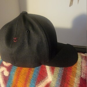 May include: A black baseball cap with a curved brim. The cap has a small red embroidered detail on the side and is resting on a colorful striped blanket. The cap is made of a textured fabric.