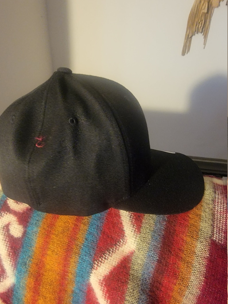 May include: A black baseball cap with a curved brim. The cap has a small red embroidered detail on the side and is resting on a colorful striped blanket. The cap is made of a textured fabric.