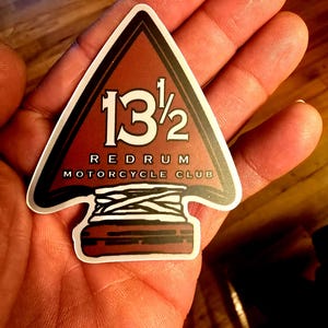May include: An arrowhead-shaped sticker with the text "13 1/2 REDRUM MOTORCYCLE CLUB." The design is mainly brown with white and black detailing. The sticker is held in a hand.