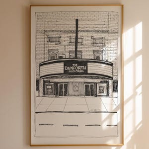 Puede incluir: Dibujo a tinta en blanco y negro de The Danforth Music Hall, enmarcado en un marco dorado. La obra de arte representa la fachada del edificio, incluyendo la marquesina con el nombre del lugar, puertas y ladrillos. El dibujo es detallado y realista.