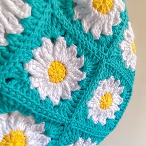 以下が含まれることがあります： デイジー模様の編み物アイテムのクローズアップ。白いデイジーの花びらに黄色の中心が、ターコイズブルーの背景に配置されています。手作りのアイテムは、テクスチャード加工された詳細なステッチを示しています。