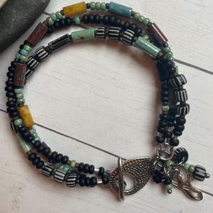 May include: A multi-strand bracelet featuring black, turquoise, yellow, and red beads. The bracelet includes black and white striped beads and a silver-colored clasp. The bracelet is designed with a mix of textures and colors.