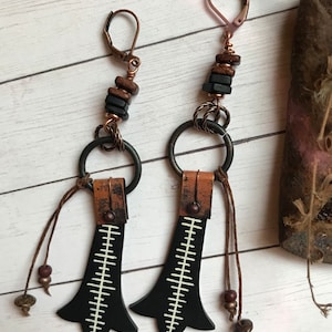 May include: A pair of black and copper earrings with leather tassels and white geometric designs. The earrings feature copper wire wrapped around black and brown beads.