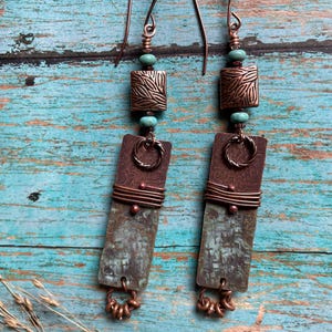 May include: Handmade copper earrings with a rustic, bohemian style. Each earring features a square, textured copper bead, turquoise beads, and a rectangular, verdigris-colored copper pendant. The earrings have a unique, artistic design with a hook closure.