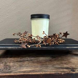 May include: A black, distressed wooden riser with a candle in a glass jar and a black lid. The candle is surrounded by a decorative arrangement of brown berries and metal star accents. The riser is on a wooden surface.