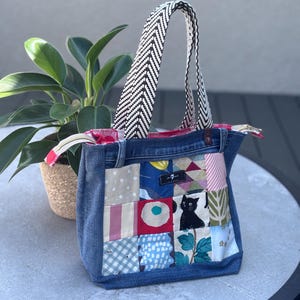 May include: A denim tote bag with a patchwork design featuring various fabric patterns and a black cat illustration. The bag has a black and white chevron patterned handle and a red and white patterned interior.
