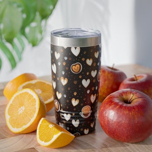 May include: A stainless steel tumbler with a black background and a pattern of brown and white hearts. The tumbler is sitting on a wooden surface with oranges and apples.