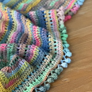 May include: A colourful crocheted shawl with a fringe border. The shawl is made with a variety of shades of blue, green, pink, and yellow yarn.