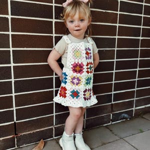 May include: A white crochet dress with colorful floral squares. The dress has a ruffled hem and suspender straps. The child is wearing white boots and has pink bows in their hair. The background is a brown brick wall.