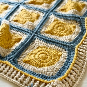 Puede incluir: Una manta de ganchillo hecha a mano con un patrón geométrico en tonos amarillo, crema y azul. La manta tiene un diseño cuadrado con un motivo en espiral en el centro de cada cuadrado. El borde es una combinación de crema y azul, con un detalle de borla.