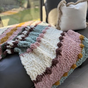 May include: A handmade, colorful blanket with a textured, plush design. The blanket features stripes of pink, white, green, and brown, with a pom-pom trim. The blanket is draped over a dark gray couch, with a white pillow in the background.