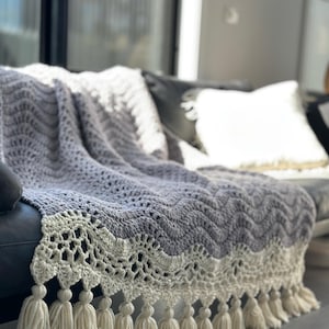 Puede incluir: Una manta de punto de ganchillo de color gris y blanco. La manta tiene un borde de ganchillo con flecos. La manta está colocada sobre un sofá.