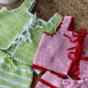 May include: Crocheted green and pink tops and shorts sets. The green set has white trim and ties, while the pink set has red trim and ties. The shorts have a striped pattern. These handmade sets are perfect for summer.