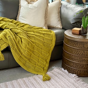 May include: A chartreuse yellow crochet throw blanket with a textured ribbed pattern and corner tassels draped over a gray leather sofa, accompanied by two cream-colored pillows and a light pink crochet rug on the floor.
