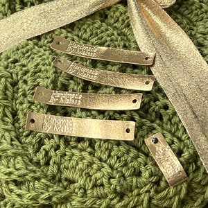 May include: Four gold-coloured leather tags with the text "Handmade by Melissa" on each tag. The tags are on top of a green crocheted blanket with a gold ribbon.