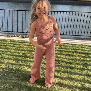 May include: A young girl smiles while wearing a handmade, dusty rose crochet jumpsuit. The jumpsuit features a V-neck top and flared pants with decorative openwork details. The girl is standing on green grass near a body of water.
