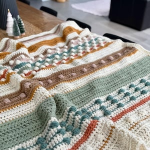 Puede incluir: Una manta de ganchillo con rayas de hilo blanco, marrón, verde, naranja y azul. La manta tiene un patrón texturizado con protuberancias elevadas y puntadas tejidas.