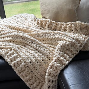 Può includere: Una coperta di lana a uncinetto color crema drappeggiata su un divano in pelle nera. La coperta ha un motivo a punto spesso e testurizzato ed è realizzata con un filato spesso.