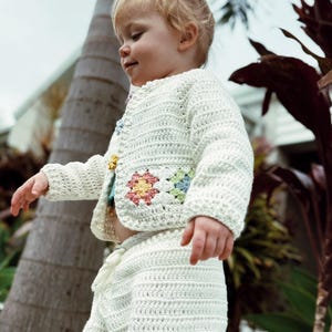 Puede incluir: Conjunto de crochet color crema para niño, con cárdigan abotonado y pantalón a juego. El cárdigan está adornado con aplicaciones florales coloridas. El conjunto está hecho de una tela texturizada de tejido abierto, perfecto para primavera o verano.