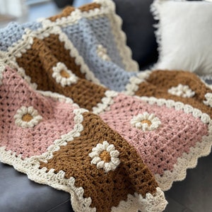 May include: A crocheted throw blanket with a patchwork design featuring brown, pink, and blue squares with white daisy accents. The blanket has a white crocheted border.