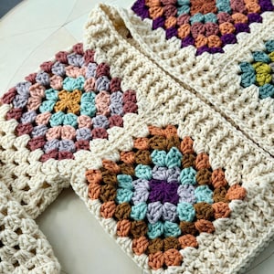 May include: A crochet cardigan constructed from cream-colored yarn, featuring multiple square motifs. Each square displays a central pattern of purple, teal, orange, and light pink yarn, framed by a thick, textured cream-colored border.