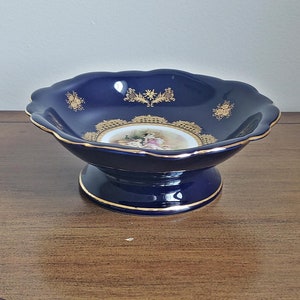 May include: A navy blue ceramic pedestal bowl with a scalloped rim and gold trim. The bowl features a central image of figures in a garden scene, surrounded by gold floral designs. A decorative piece for display.