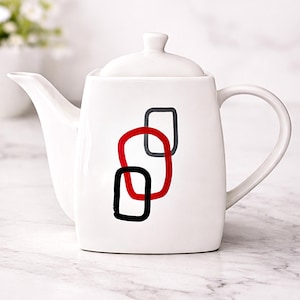 May include: White ceramic teapot with a lid and handle. The teapot features a design of three overlapping geometric shapes in red, black, and gray. The teapot is on a white marble surface.