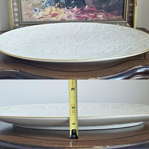 May include: A white oval serving platter with a gold rim and an embossed floral design. The platter is sitting on a wooden table.