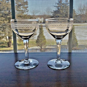 May include: Two clear, stemmed glasses with etched decorative patterns near the rim. The glasses sit on a dark surface in front of a window with a view of trees and a screen.
