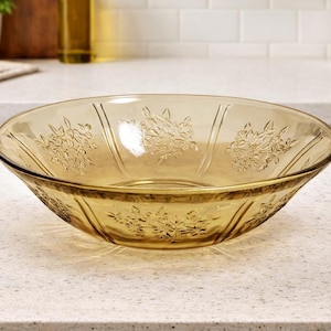 May include: Amber-colored glass bowl with floral embossed design. The bowl has a wide, shallow shape and is sitting on a white countertop. The kitchenware is likely used for serving salads or other dishes.