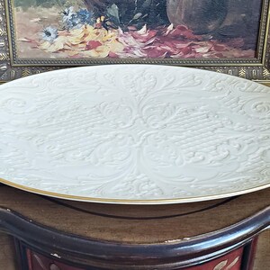 May include: A large, white, ceramic serving platter with an embossed, floral design. The platter has a gold rim.