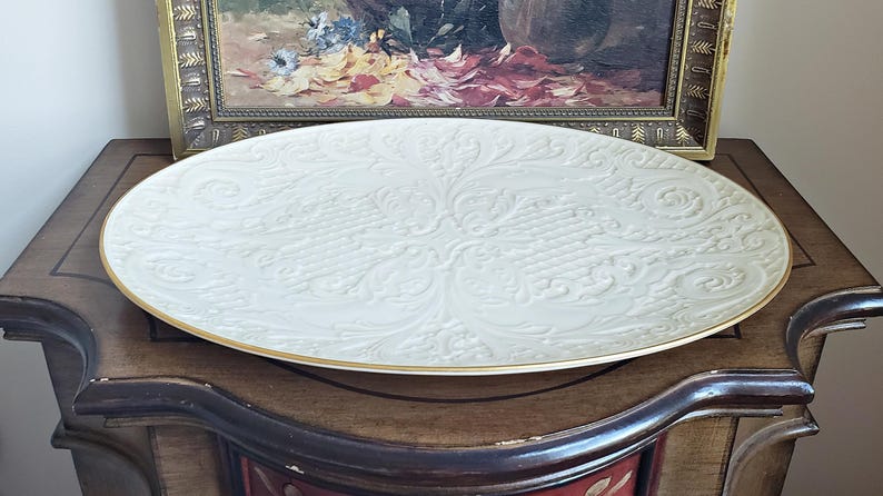 May include: A large, white, ceramic serving platter with an embossed, floral design. The platter has a gold rim.