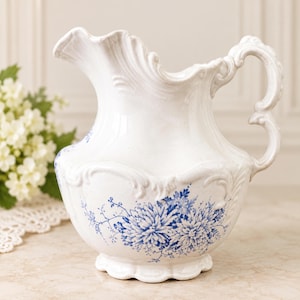 May include: A white ceramic jug featuring an ornate, scalloped rim and a curved handle. The body of the jug is decorated with a detailed blue floral pattern of chrysanthemums and delicate foliage, set against a glossy white finish.