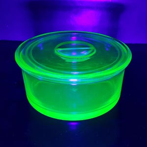 May include: A glowing, neon green glass bowl with a matching lid. The bowl is round with a slightly flared rim and a clear handle on the lid. The glass appears to be vintage and glows brightly under a black light.