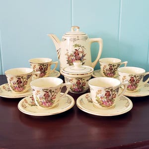 Puede incluir: Un juego de té vintage color crema con ribetes dorados, que incluye una tetera, azucarero y seis tazas con platillos. Cada pieza está decorada con una colorida escena pastoral. El juego está dispuesto sobre una superficie de madera oscura.