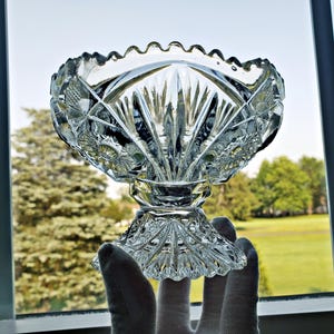 Könnte beinhalten: Eine transparente, geschliffene Glasschale mit gewelltem Rand und Sockel. Die Schale weist komplizierte geometrische und florale Muster auf, die das Licht reflektieren. Die Schale wird vor einem Fenster mit Blick auf Bäume und einen hellen Himmel gehalten.