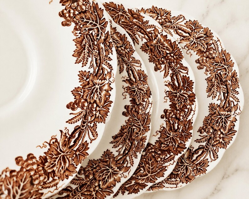 May include: A set of four white ceramic plates with scalloped edges, each featuring a detailed brown transferware pattern of grapevines, leaves, and clusters of grapes encircling the rim in a continuous, intricate botanical design.