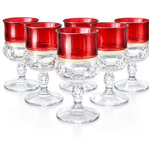 May include: Six clear crystal wine glasses with ruby red bowls and gold trim. The glasses have a decorative, cut-glass design on the stems and bowls. The glasses are arranged in a triangular formation against a white background.