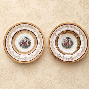 May include: Two round porcelain plates featuring a central circular medallion with a colorful illustration of figures in a landscape. The plates have a wide white rim decorated with intricate gold filigree patterns and a deep red border.