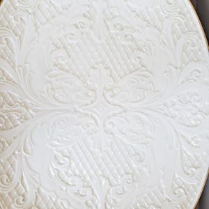 May include: A white ceramic plate with a raised, embossed design of intricate floral patterns. The plate has a gold rim.
