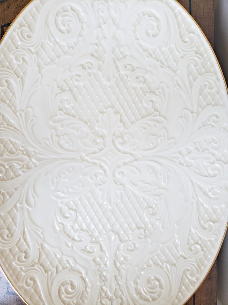May include: A white ceramic plate with a raised, embossed design of intricate floral patterns. The plate has a gold rim.