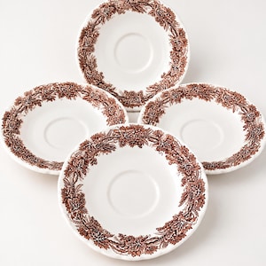 Peut inclure: Un ensemble de quatre soucoupes en céramique blanche, chacune ornée d'un motif brun imprimé sur le bord. Le design se compose d'illustrations détaillées de grappes de raisin et de feuilles de vigne disposées en une bordure circulaire continue.